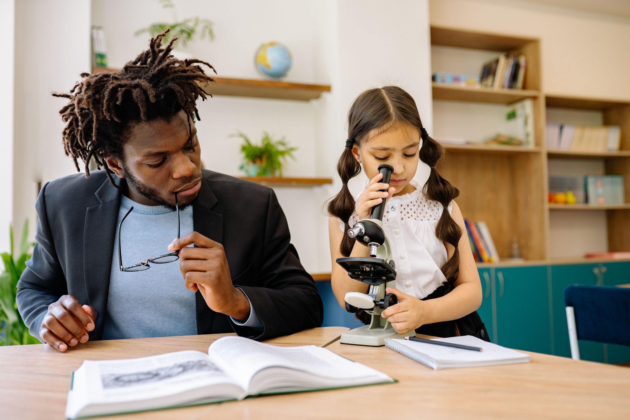A teacher guiding a child as she looks through a microscope.